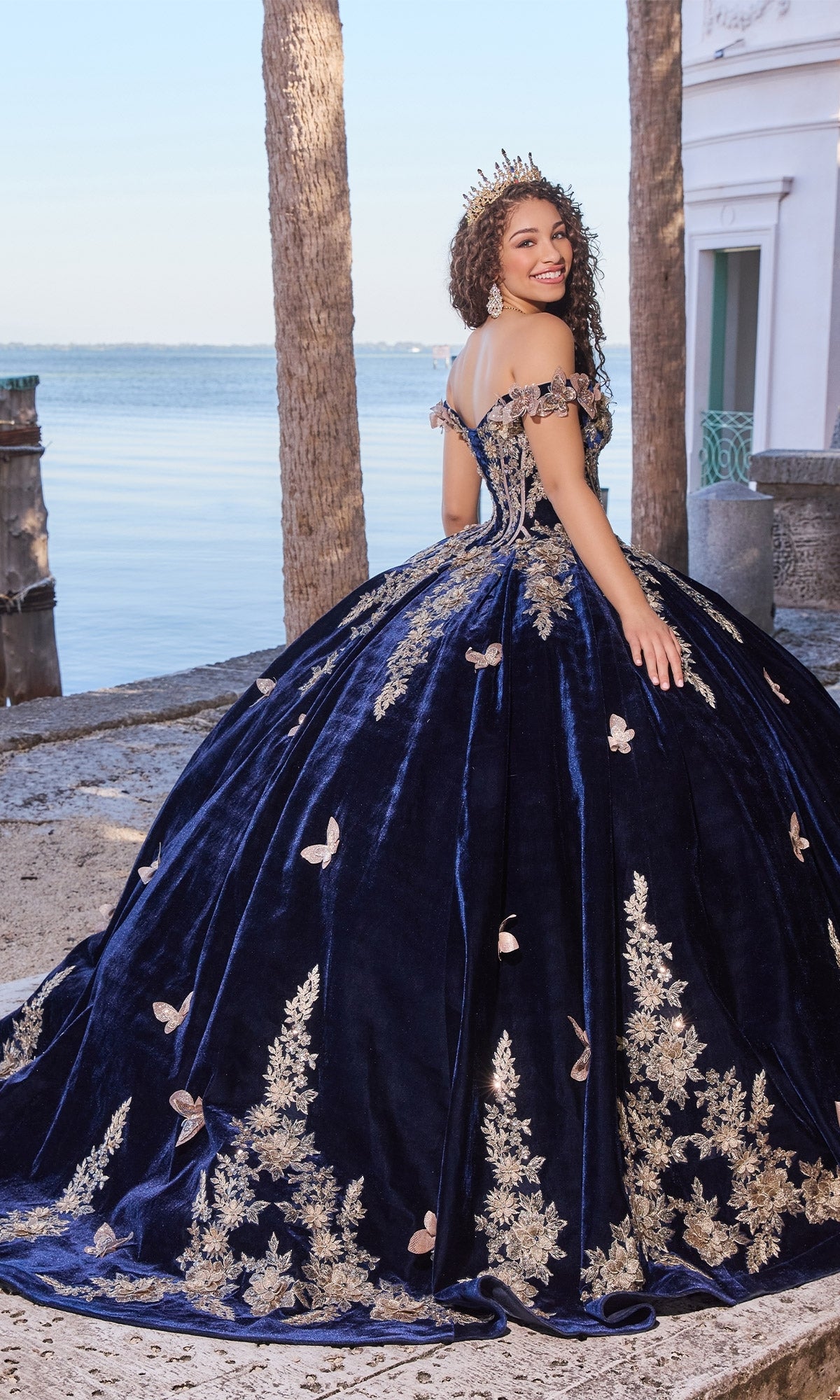 A young woman smiles outdoors by the water, wearing the Velvet Quinceanera Dress: Princesa PR30136 in navy blue velvet with gold lace embroidery, butterfly details, and a crown.